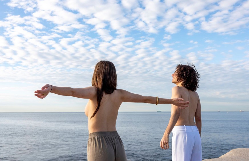 Eine Frau steht oberkörperfrei mit ihrem Freund am Meer und genießt die Freiheit.