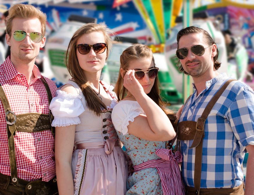 Group of happy friends celebrating Beer Fest posing in front of a carousel wearing traditional tracht and sunglasses