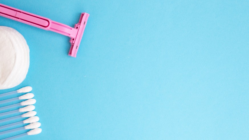 top view personal care products. white bottle, pink razor, ear sticks, cotton pads, toothbrush on blue background. copy space