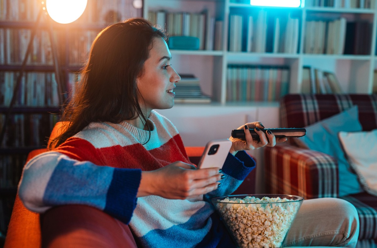 Young woman relaxing at home, eating popcorn and using phone while watching TV