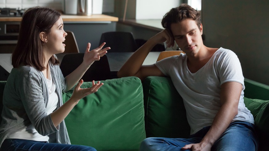 Young attractive couple sitting on couch at home and sorting out their relations. Girlfriend talking too much, drama queen nagging disinterested boyfriend feels angry. Troubles and break up concept