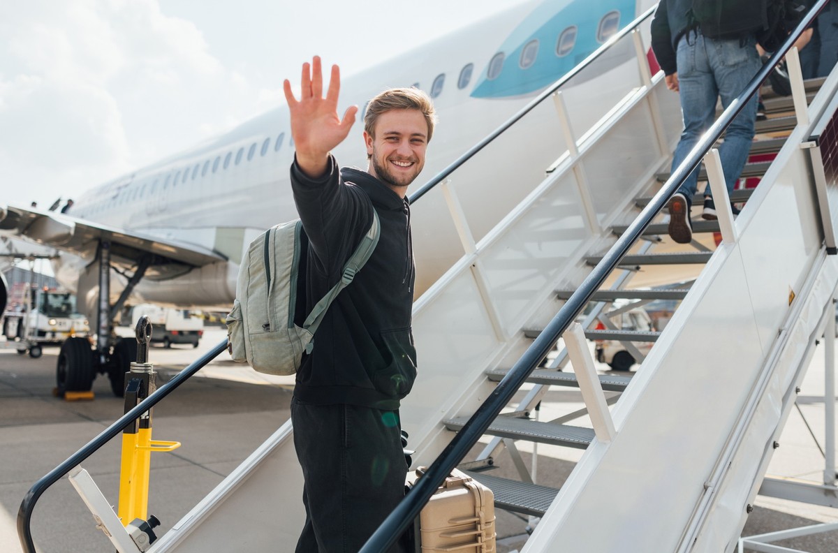 A young backpacker getting onto a plane while budget travelling. He is walking up the steps to the plane carrying his suitcase, smiling at the camera while waving.