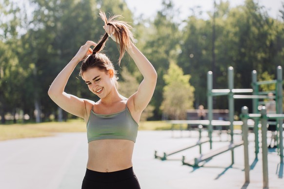 Excited Spanish girl in sportswear standing at park gathering hair in ponytail against sports ground, laughing dreaming. Fit ambitious fashion model at workout. Healthy lifestyle, fitness. Active.