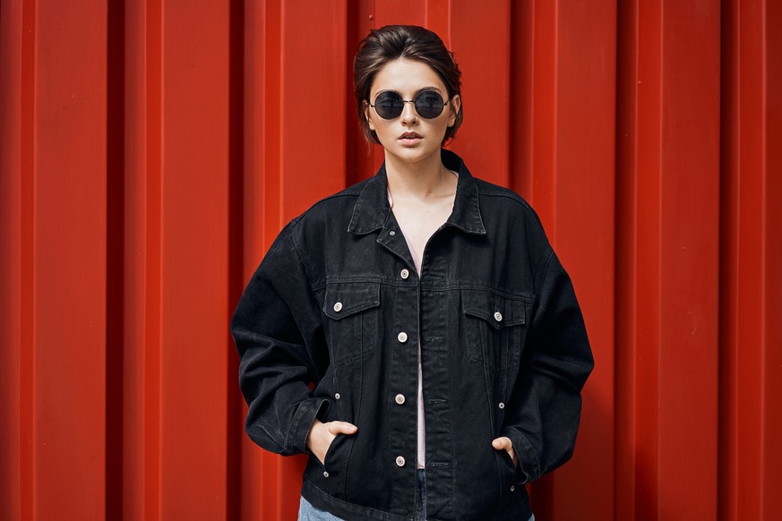 Young hipster woman wear sunglasses and black denim jacket isolated on red background