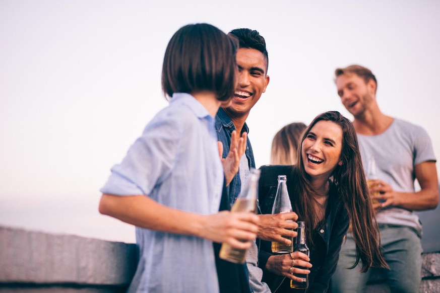 Young adult multi-ethnical friends hanging out on rooftop after work to drink together laughing and joking