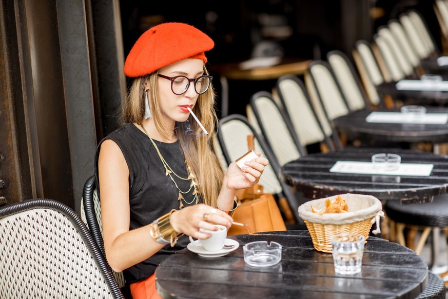 Young stylish woman in red beret smoking a cigarette sitting outdoors at the french cafe