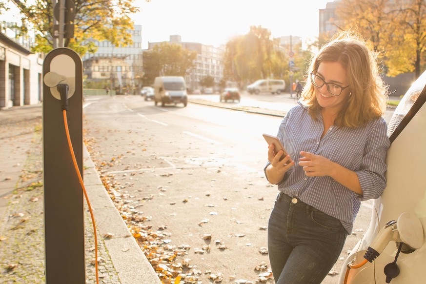 Young woman is standing near the electric car and holding smartphone. The rental car is charging at the charging station for electric vehicles. Car sharing.