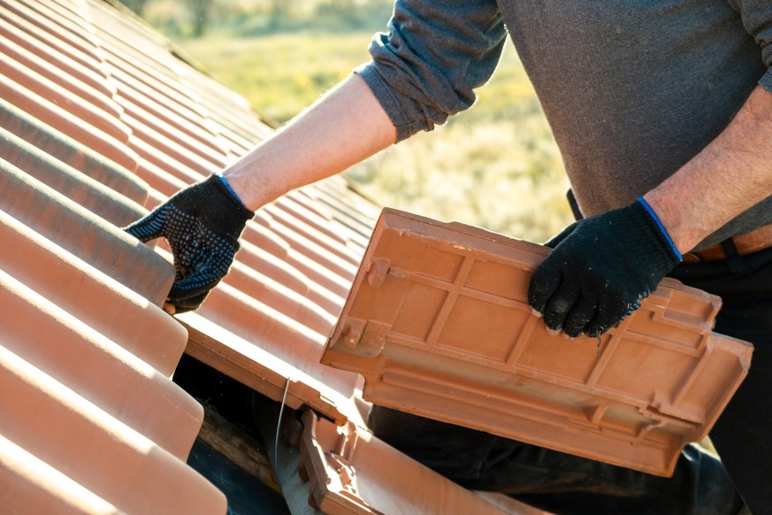 Closeup of worker hands installing yellow ceramic roofing tiles mounted on wooden boards covering residential building roof under construction.