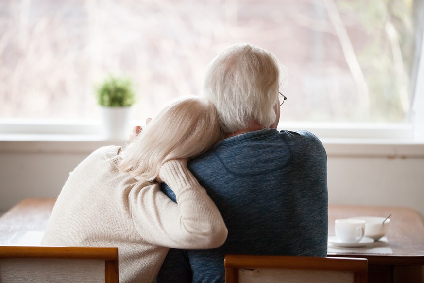 Rear view at senior grey haired loving caring family couple embracing relaxing at home together enjoying peaceful morning breakfast looking at window view thinking of future feeling calm nostalgic