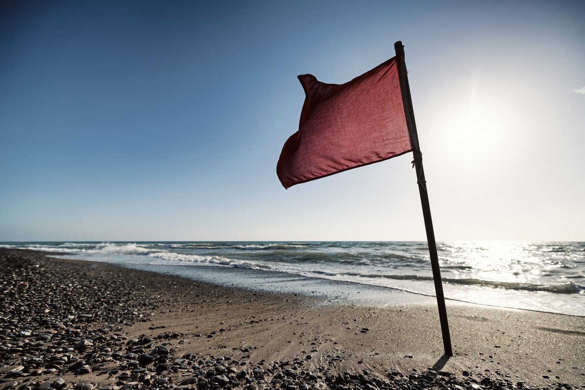 A red flag stands on a sandy beach, indicating hazardous swimming conditions. Sea waves crash against the shore under a clear blue sky, with the sun shining brightly in the background. The summer sea ...