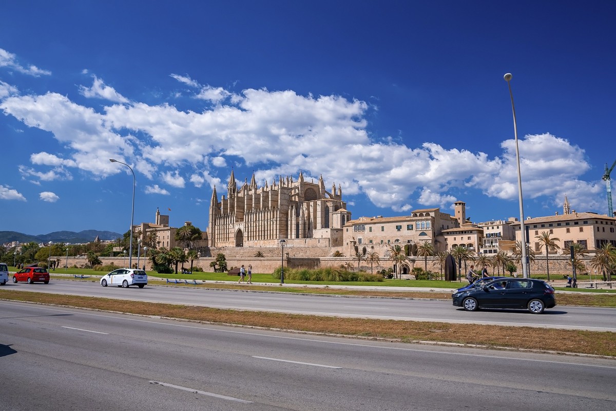 Mallorca, Spain. April 27, 2022. Cars moving on city street by beautiful gothic La Seu Cathedral. Vehicles on road against blue sky. Famous tourist attraction in historic old town.