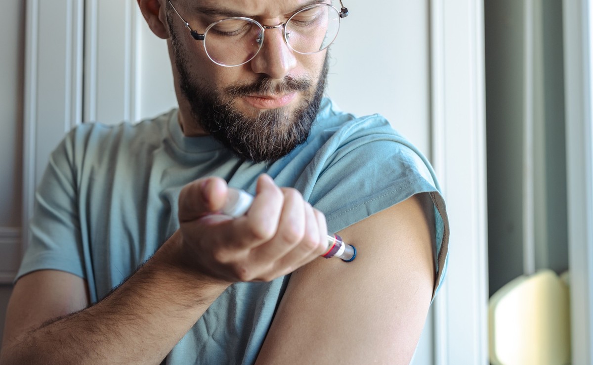 A man is using an injection device on his arm in a brightly lit room, emphasizing personal healthcare and responsibility.