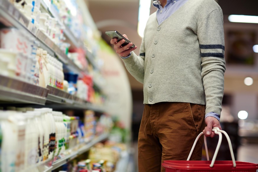 Mid-section portrait of unrecognizable man using smartphone for shopping list, choosing dairy products in supermarket