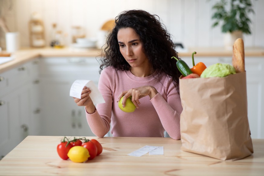 Concerned Young Woman Checking Bills In Kitchen After Grocery Shopping, Millennial Female Sitting At Table In Kitchen, Frustrated About Expensive Vegetables And Fruits, Closeup Shot, Selective Focus