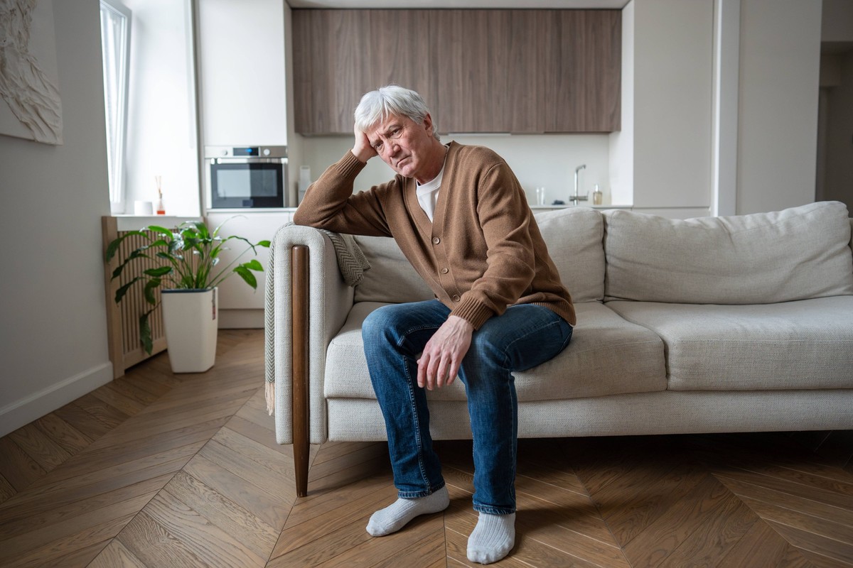 Depressiver, apathischer alter Mann, der den Kopf in die Hand stützt und allein auf der Couch sitzt, mit leerem Blick zu Hause Lonely senior sits motionless supporting heavy head with hand showing adv ...