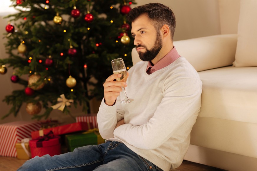 Lonely Christmas. Sad young man sitting alone on the floor near a Christmas tree and drinking champagne, while looking disappointed
