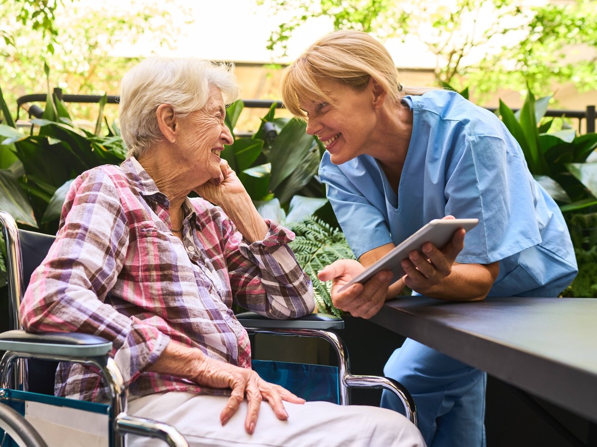 A nurse cheerfully interacts with a senior in a wheelchair, using a tablet to communicate effectively, in a serene outdoor setting with lush greenery, indicating care, companionship, and technology in ...