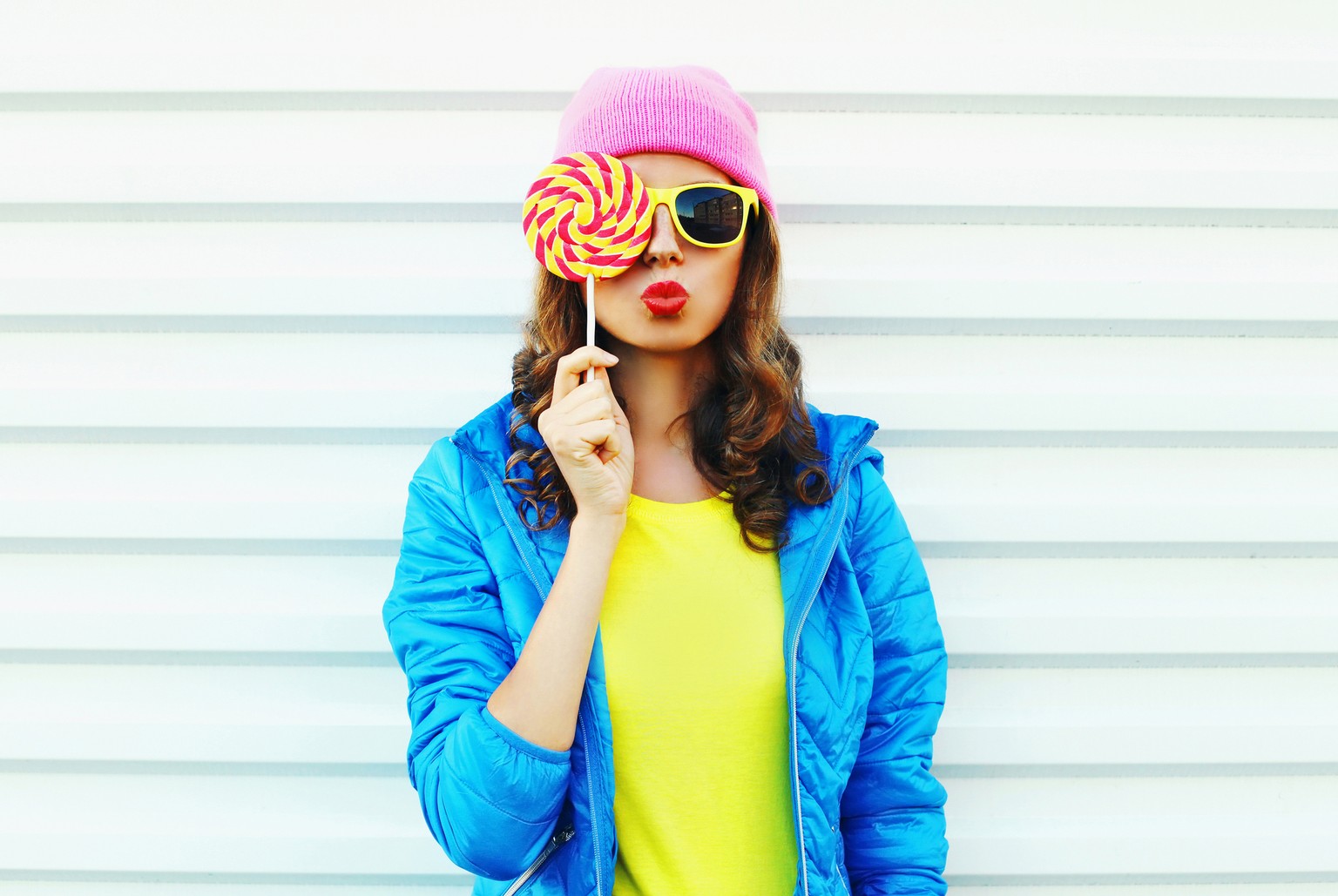 Portrait fashion pretty cool woman with lollipop in colorful clothes over white background wearing a pink hat yellow sunglasses and blue jacket