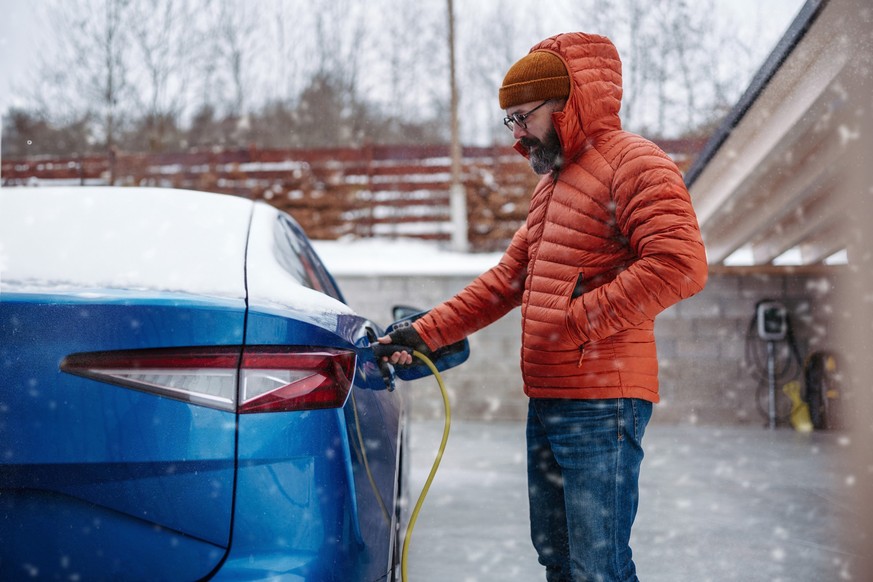 Man charging electric car during cold snowy day. Side view of hansome mature man putting charger in charging port during cold weather. Charging and driving electric vehicles during winter season.