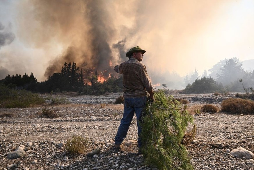 A local man holds a pine tree branch to beat down the approaching wildfires burn in the background, near the village of Vati, just north of the coastal town of Gennadi, in the southern part of the Gre ...