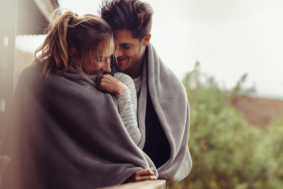 Romantic couple on a winter holiday. Man and woman standing together in a hotel room balcony wrapped in blanket. Couple embracing and smiling.