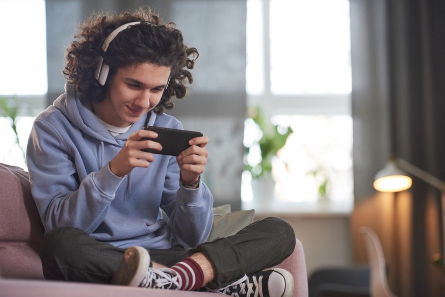 Teenage boy in wireless headphones sitting on sofa using his mobile phone for playing online games