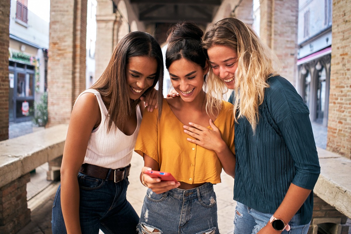 Three happy women using mobile phone outdoors. Group of smiling female friends watching social media at smartphone. High quality photo