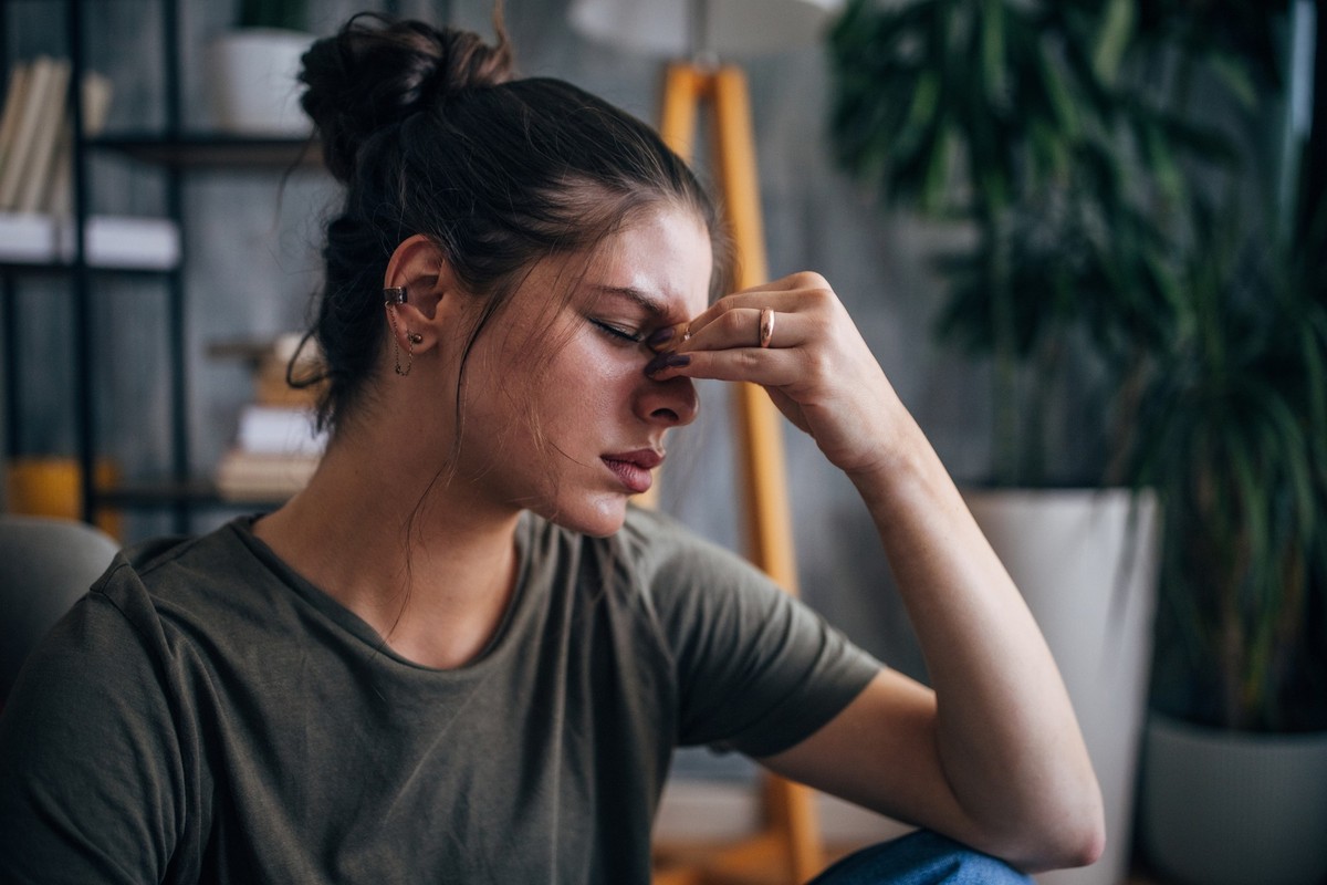 The woman, sitting in a cozy living room with plants and books in the background, touches her forehead, indicating that she is feeling uncomfortable or stressed.