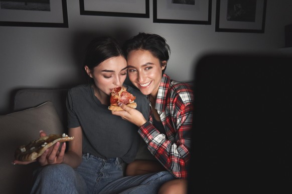 Two happy young girls eating pizza and watching tv at home party late. Best friends or couple having fun enjoying movie on television streaming service eating at night sitting on couch together.