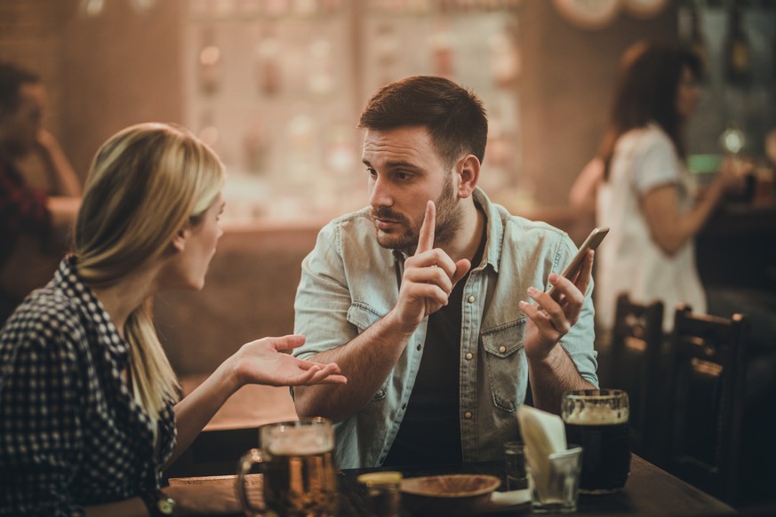 Young man using cell phone in a bar and arguing with his girlfriend.