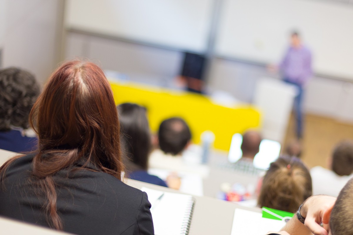 Speaker giving presentation in lecture hall at university. Participants listening to lecture and making notes.