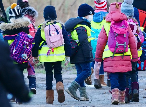 28.01.2022, Berlin. Schüler einer ersten Klasse einer Grundschule folgen bei einem Klassenausflug an einem Januartag ihrer Lehrerin. Dabei tragen sie Rucksäcke mit Proviant und gelbe Warnwesten. Ein J ...