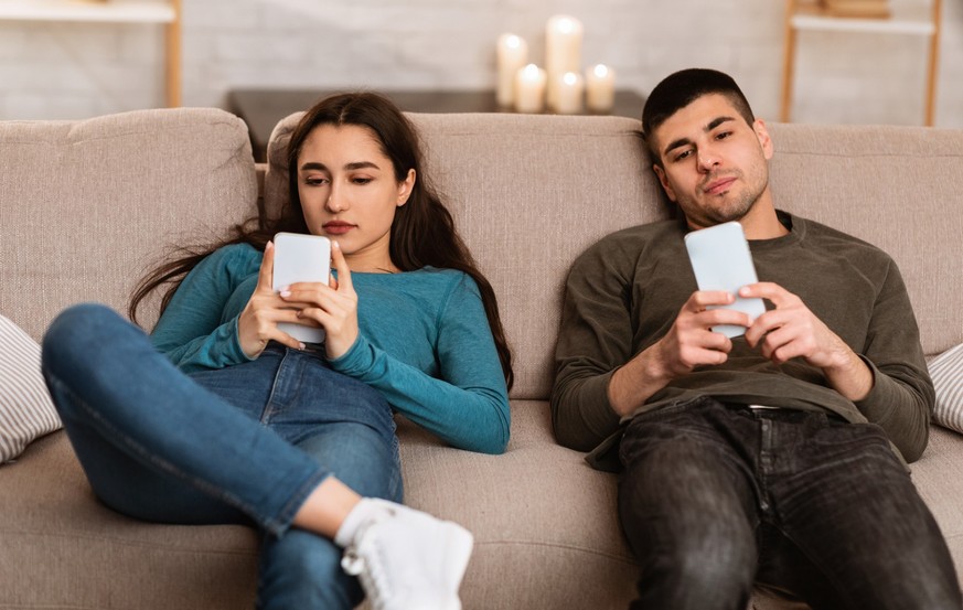 Boredom Concept. Young couple using smart phones, sitting on couch at home during self isolation