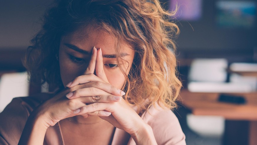 Closeup of sad young Asian woman at cafe leaning head on clasped hands and staring into vacancy. Tired freelancer feeling burnout. Stress and bad news concept