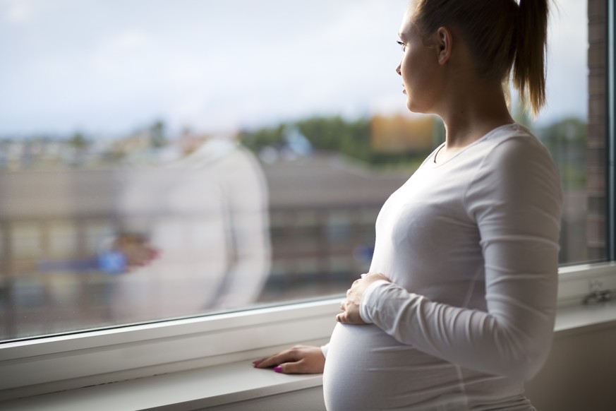 Thoughtful pregnant woman looks out the window home. Holding a hand on the tummy. Mental health and pregnancy.