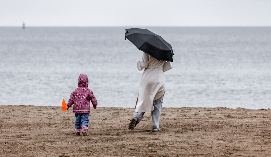 29.03.2024, Schleswig-Holstein, Travemünde: Eine Frau mit Schirm und ein Kind sind bei diesig trübem Wetter am Ostsee-Strand in Travemünde unterwegs. (zu dpa: «Aprilwetter bringt Dauerregen in den Nor ...