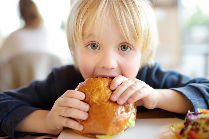 Cute blonde boy eating large hamburger at fast food restaurant. Unhealthy meal for kids. Junk food. Overweight problem child.