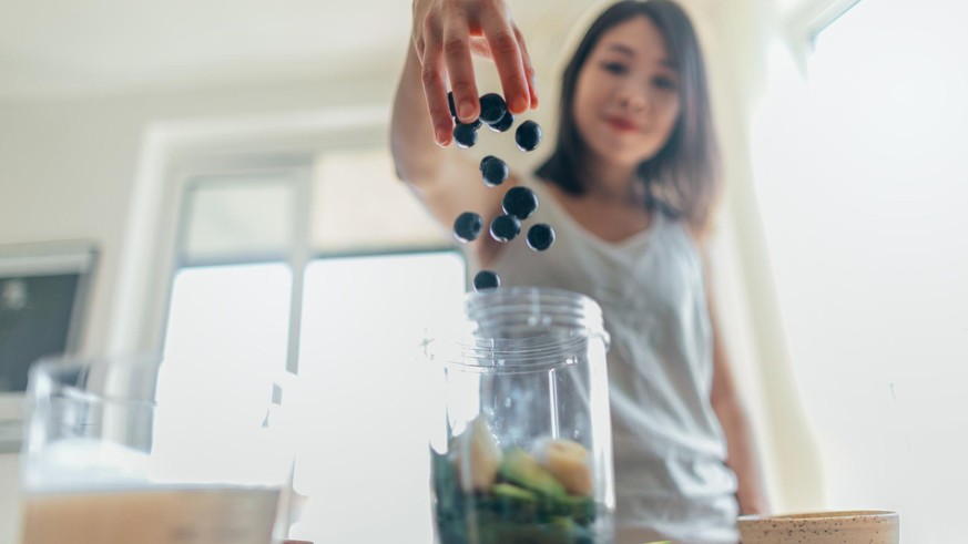 Low angle view of a beautiful young Asian woman throwing blueberry into the blender to make a healthy smoothie with banana, spinach and vegan milk.