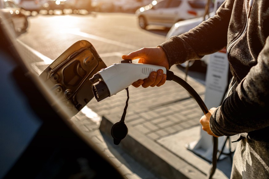 Man Holding Power Charging Cable For Electric Car In Outdoor Car Park. And heâs going to connect the car to the charging station in the parking lot near the shopping center.