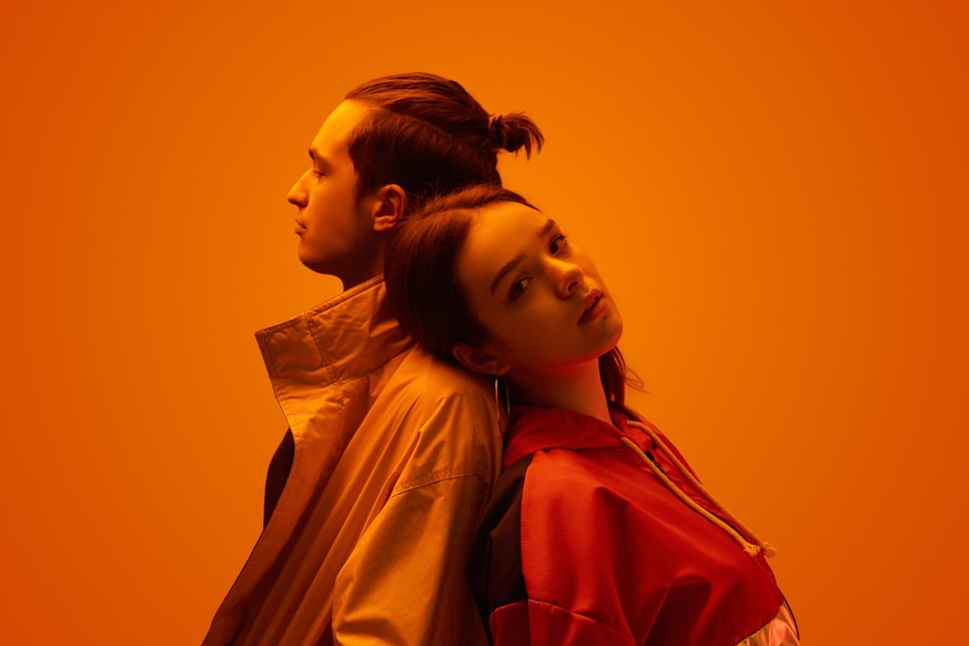 Side view of young couple of teen boyfriend and girlfriend in trendy clothes, standing back to back on orange background in studio with neon light love