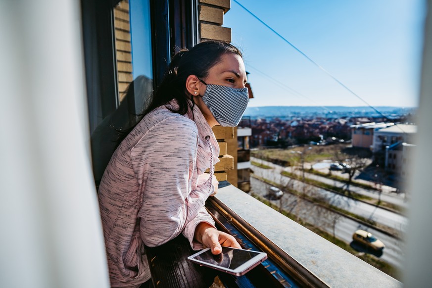 Woman in quarantine with face mask using smartphone and looking through the window. Pandemic of Coronavirus / Covid-19.