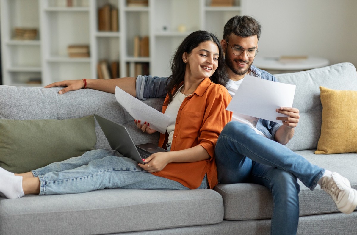 Happy couple checking documents, spouses reading insurance agreement or property certificate, husband and wife sitting on couch in living room, copy space
