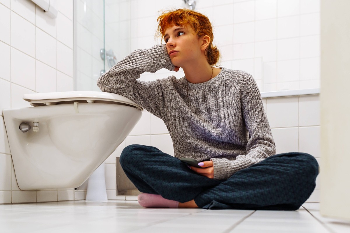 Porträt Teenager-Mädchen sitzt in der Toilette auf dem Boden mit Telefon in der Hand portrait red-haired, tired, depressed teenage girl sitting on floor in bathroom, resting her head on hand, leaning ...