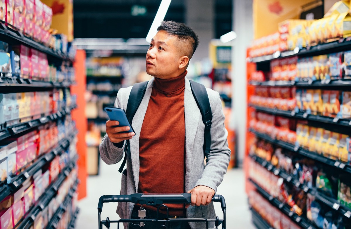 Handsome Asian male walking down the product aisle in the supermarket, looking at shelves and searching for groceries from the list on his mobile phone he is holding in his hand.