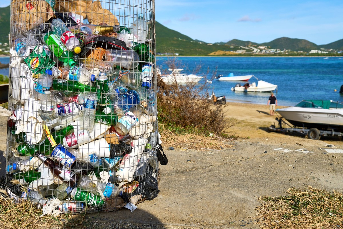 Sint Maarten, Caribbean - January 3, 2025: An overfilled wire garbage can full of plastic waste stands on the coast of Sint Maarten. Boats lie on the beach in the background. A contrast between nature ...