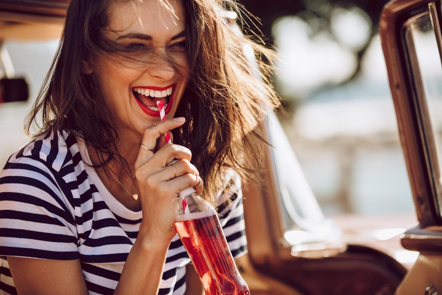 Closeup of beautiful young woman enjoying drinking cola with straw while sitting in a car. Female on a road trip having soft drink.