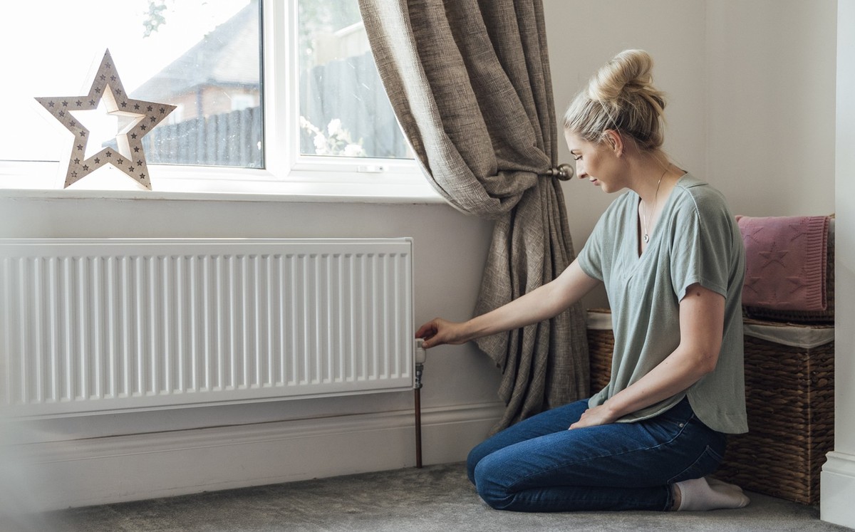 Side view of a young woman kneeling on the living room floor, adjusting the thermostat on the radiator.