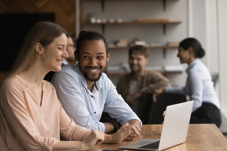 Multi ethnic workmates Caucasian woman African man talk laugh sit in coworking space, working together use laptop enjoy communication. Teamwork, friendly good work relations, cooperation, fun concept