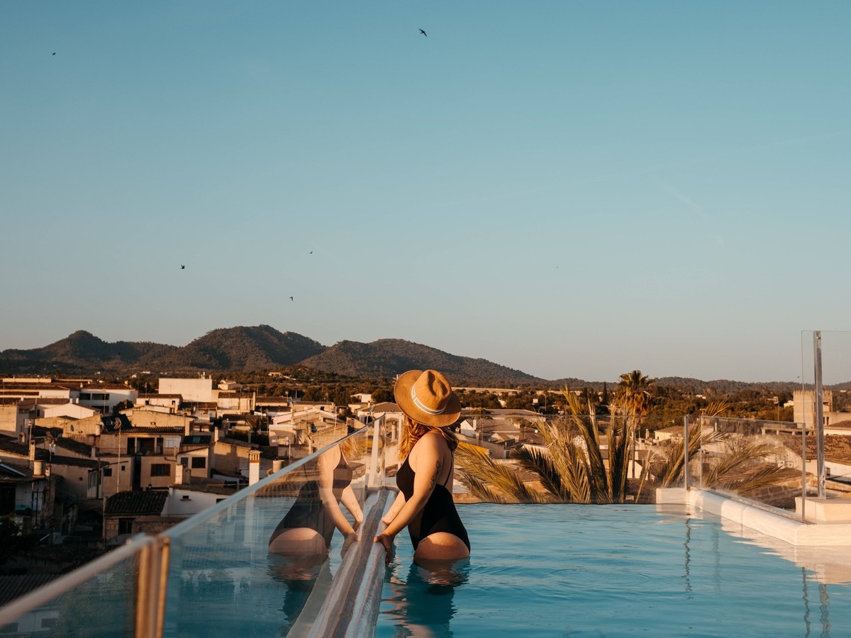 Side view of unrecognizable young female tourist in stylish black swimsuit and straw hat standing in swimming pool on rooftop and admiring town against cloudless blue sky in Mallorca, Model released E ...