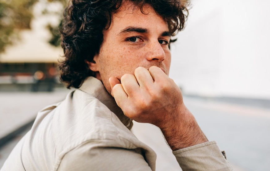 Portrait of serious young man with curly hair looking at the camera, sitting outside. Portrait of handsome male with curly hair has serious expression, take a rest in the city street.